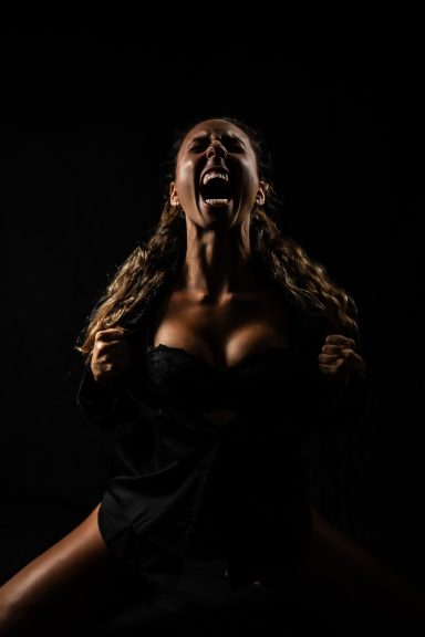 Mujer con cabello rizado, gritando en un entorno oscuro, vistiendo ropa negra.
