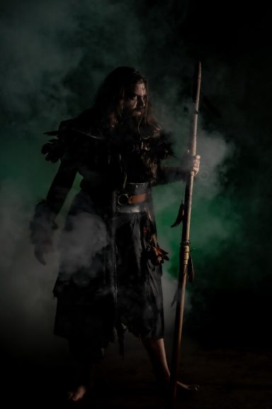 Personaje con lanza en un ambiente oscuro y brumoso, vestido con ropas místicas.