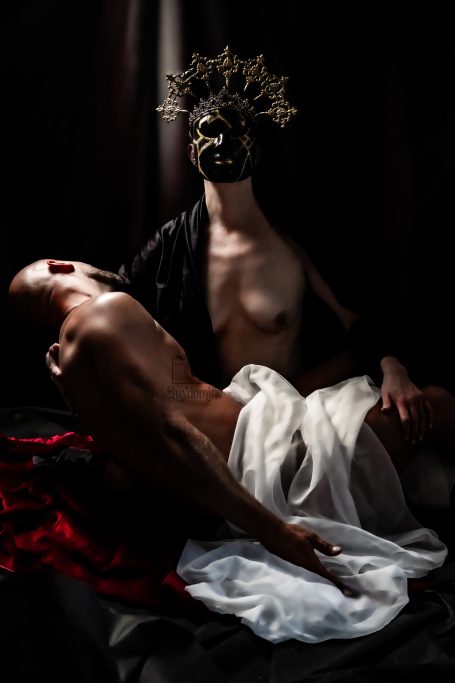 La piedad Figura masculina reclinada con una persona enmascarada sobre él, en un entorno oscuro.