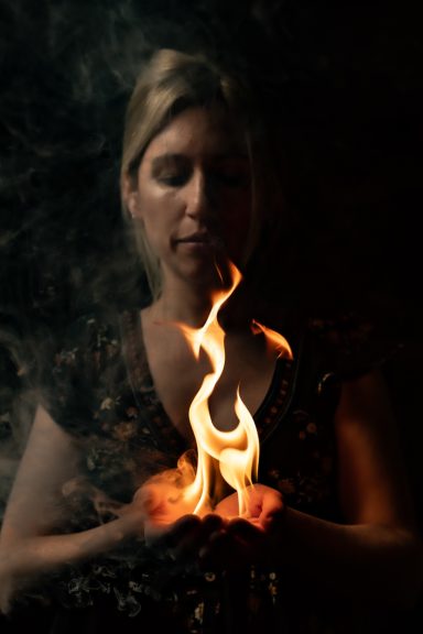 Mujer sosteniendo fuego entre sus manos, con fondo oscuro y humo.