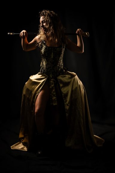 Mujer con vestido elegante sosteniendo una vara, con fondo oscuro.