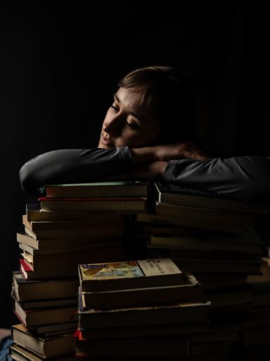 Persona pensativa apoyada sobre una pila de libros en un entorno oscuro.