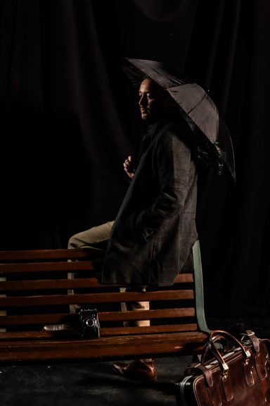 Persona sentada en un banco, sosteniendo un paraguas, con un maletín junto a ella.