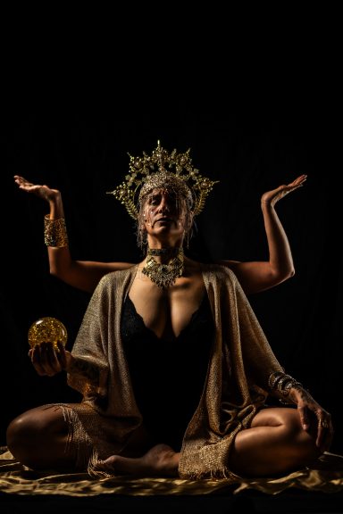 Mujer en postura de meditación con corona, sosteniendo un cráneo y brazos elevados.