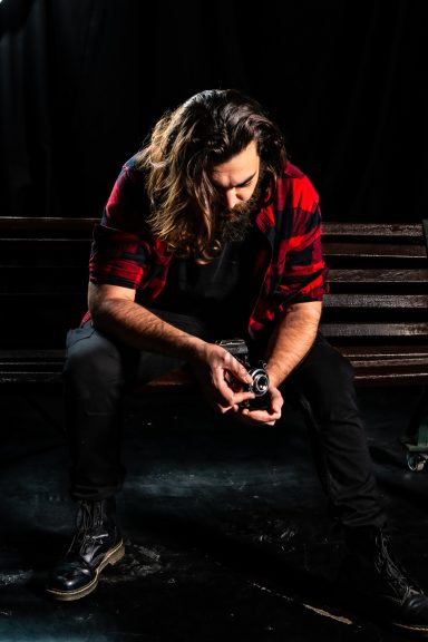Hombre con cabello largo y barba, vistiendo una camisa de cuadros, sentado y mirando un objeto.
